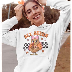 Pu&ograve; includere: Felpa con cappuccio bianca con una grafica di un tacchino dei cartoni animati con un fiocco rosa. Il testo "SIX SEVEN" &egrave; sopra il tacchino, con stelle e foglie autunnali. Un motivo a scacchi in bianco e nero &egrave; sotto.