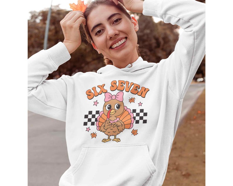 Pu&ograve; includere: Felpa con cappuccio bianca con una grafica di un tacchino dei cartoni animati con un fiocco rosa. Il testo "SIX SEVEN" &egrave; sopra il tacchino, con stelle e foglie autunnali. Un motivo a scacchi in bianco e nero &egrave; sotto.