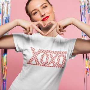 Pu&ograve; includere: Una maglietta bianca con un motivo a scacchi rosso e bianco che dice "XOXO" con una fila di piccoli cuori rossi sotto il testo.
