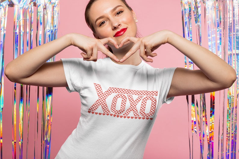 Pu&ograve; includere: Una maglietta bianca con un motivo a scacchi rosso e bianco che dice "XOXO" con una fila di piccoli cuori rossi sotto il testo.
