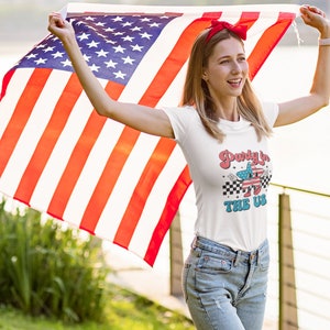 May include: A woman wearing a white t-shirt with the text "Party in the US" and a red headband holds an American flag in front of her.