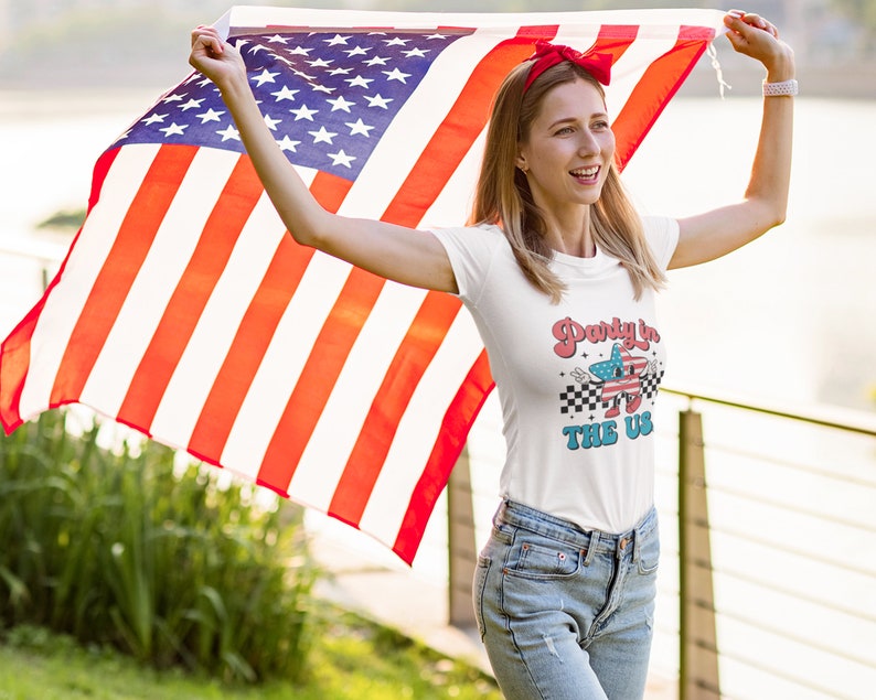 May include: A woman wearing a white t-shirt with the text "Party in the US" and a red headband holds an American flag in front of her.