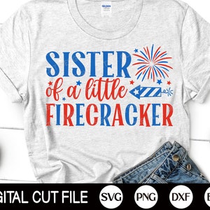 May include: A gray t-shirt with the text "Sister of a little Firecracker" in red and blue lettering. A red, white, and blue firework graphic is above the text. The shirt is on a white surface with a pair of blue jeans and a pair of red sunglasses.