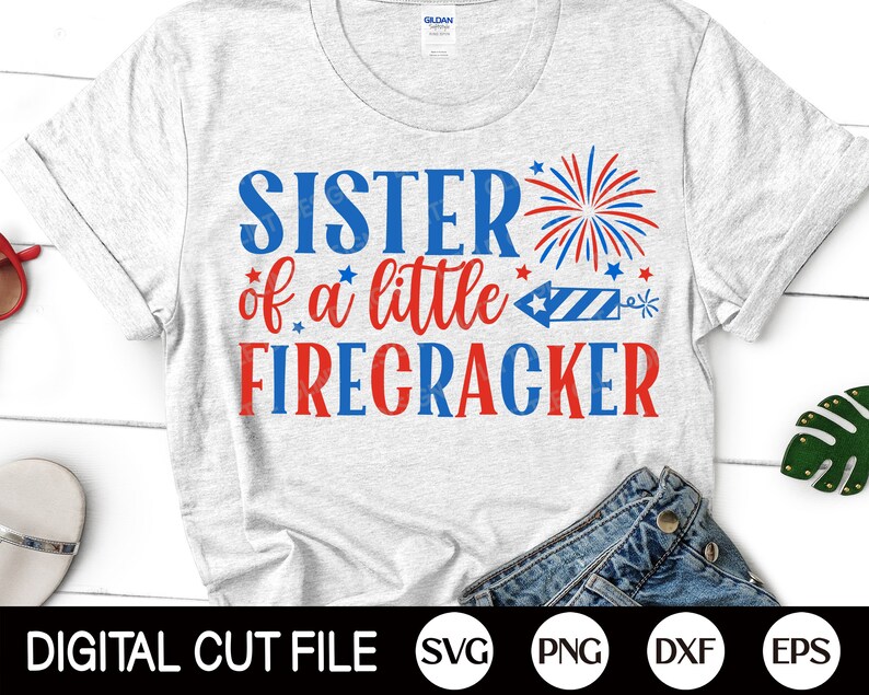 May include: A gray t-shirt with the text "Sister of a little Firecracker" in red and blue lettering. A red, white, and blue firework graphic is above the text. The shirt is on a white surface with a pair of blue jeans and a pair of red sunglasses.