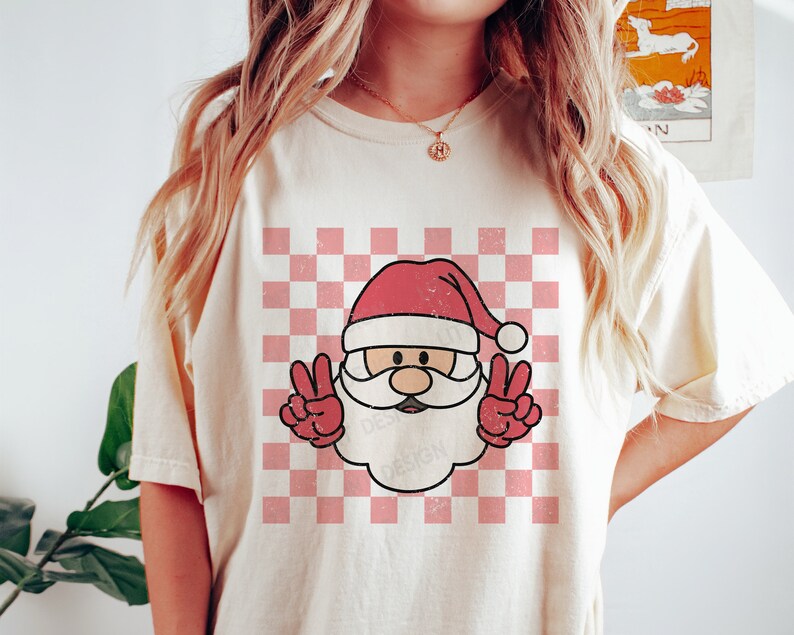 May include: A white t-shirt with a pink and white checkered pattern and a cartoon image of Santa Claus with a red hat and beard, giving the peace sign with both hands.