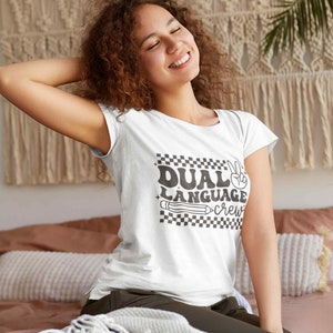 May include: White t-shirt with a black and white checkered design and the text "Dual Language Crew" in a retro font. A peace sign and a pencil are also included in the design.