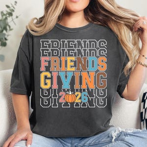 May include: A charcoal gray t-shirt featuring the text FRIENDS GIVING in a stacked, multi-colored block font. The design includes a small orange pumpkin graphic replacing the letter O in the year 2026, centered below the main text.
