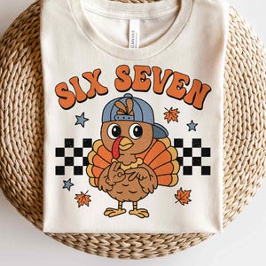 Pu&ograve; includere: T-shirt color crema con un tacchino dei cartoni animati con un berretto blu. Il tacchino ha piume arancioni e marroni e fa il segno della pace. Il testo "SIX SEVEN" &egrave; in arancione, con un lettering in stile retr&ograve; sopra il tacchino. Motivo a scacchiera in bianco e nero in basso.
