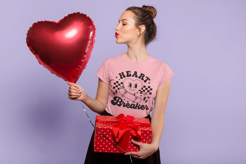 K&ouml;nnte beinhalten: Eine Frau tr&auml;gt ein pinkfarbenes T-Shirt mit dem Text "HEART BREAKER" und einem Comic-Herz auf einem Skateboard. Sie h&auml;lt einen roten herzf&ouml;rmigen Ballon und eine rote Geschenkbox mit wei&szlig;en Punkten.