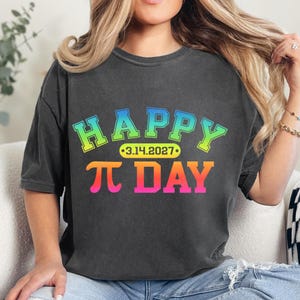 Pu&ograve; includere: T-shirt grigio scuro con le parole "HAPPY &pi; DAY" in un design a gradiente colorato. Il testo include la data "3.14.2027" in un ovale giallo. La maglietta &egrave; in stile casual.