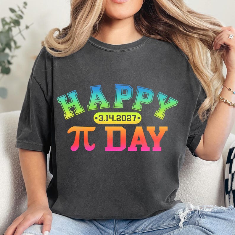 Pu&ograve; includere: T-shirt grigio scuro con le parole "HAPPY &pi; DAY" in un design a gradiente colorato. Il testo include la data "3.14.2027" in un ovale giallo. La maglietta &egrave; in stile casual.