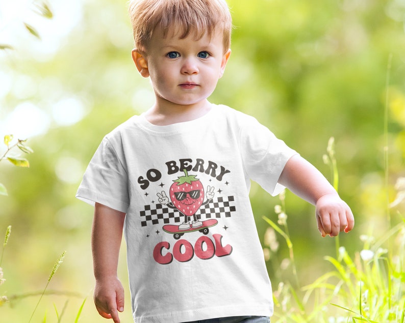 K&ouml;nnte beinhalten: Ein wei&szlig;es T-Shirt mit einer Comic-Erdbeere, die eine Sonnenbrille tr&auml;gt und Skateboard f&auml;hrt. Der Text "SO BERRY COOL" ist auf das Shirt gedruckt.