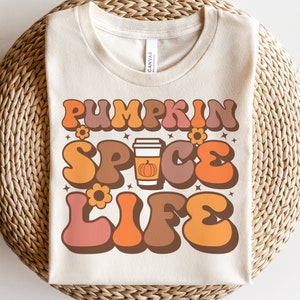 May include: A white t-shirt with a retro-style graphic that says "Pumpkin Spice Life" in orange, brown, and pink letters. A stylized image of a pumpkin spice latte is in the center of the graphic.