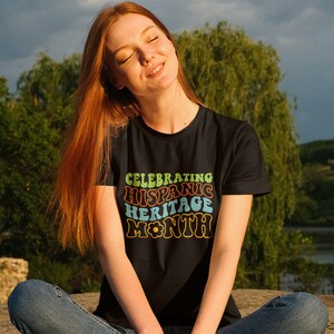 May include: A woman wearing a black t-shirt with a retro-style graphic that says "Celebrating Hispanic Heritage Month".