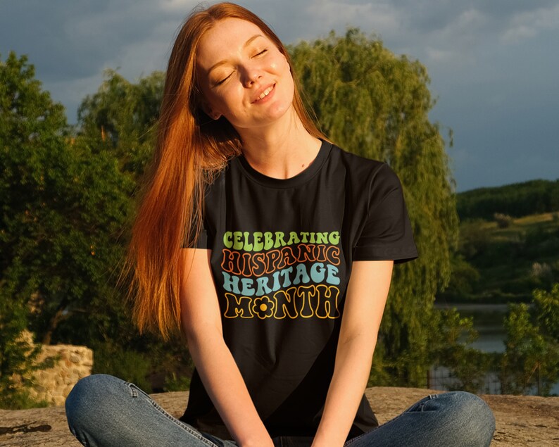 May include: A woman wearing a black t-shirt with a retro-style graphic that says "Celebrating Hispanic Heritage Month".