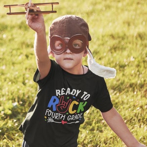 May include: A child wearing a brown aviator hat and goggles is holding a toy airplane. The child is wearing a black t-shirt with the text "Ready to Rock Fourth Grade" in colorful letters.