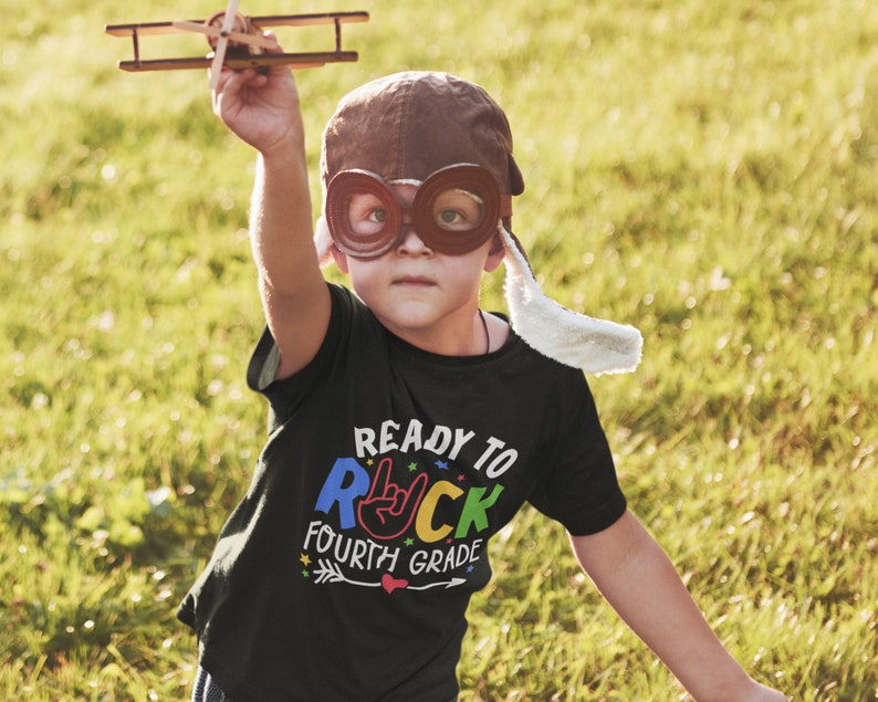 May include: A child wearing a brown aviator hat and goggles is holding a toy airplane. The child is wearing a black t-shirt with the text "Ready to Rock Fourth Grade" in colorful letters.