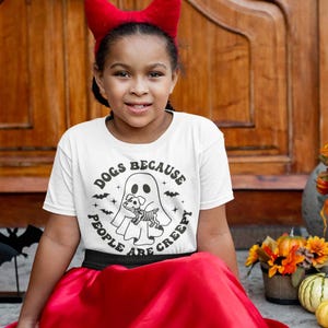 May include: A young person wearing a white t-shirt with a black and white graphic of a ghost holding a dog. The text on the shirt reads "Dogs because people are creepy". The person is wearing a red devil horn headband and a red skirt.