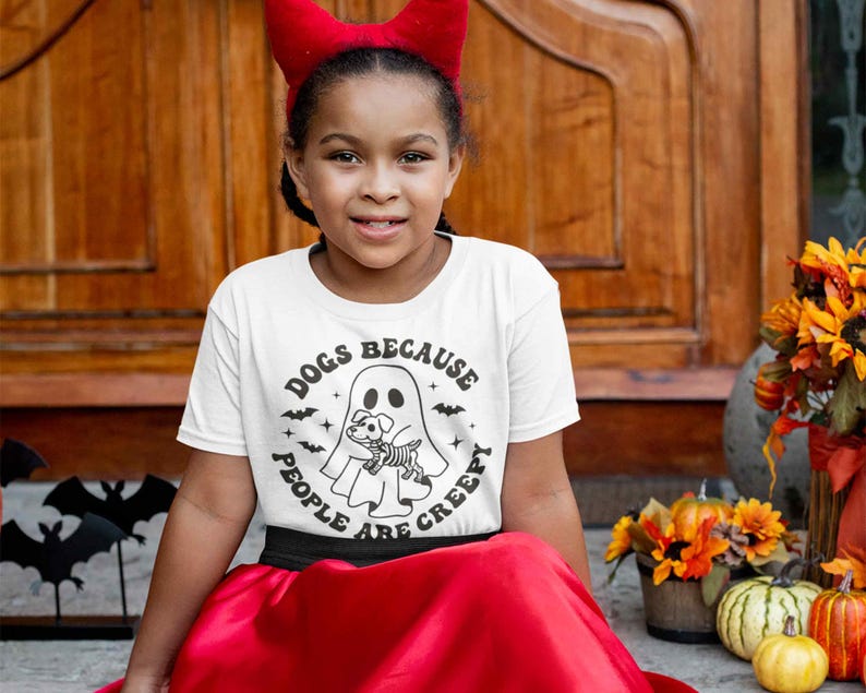 May include: A young person wearing a white t-shirt with a black and white graphic of a ghost holding a dog. The text on the shirt reads "Dogs because people are creepy". The person is wearing a red devil horn headband and a red skirt.