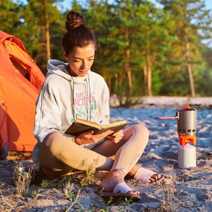 Pode incluir: Uma pessoa usando um moletom branco com a frase "LIFE IS BETTER" lendo um livro ao ar livre. Uma barraca laranja e um fog&atilde;o de acampamento s&atilde;o vis&iacute;veis ao fundo. A pessoa est&aacute; sentada na areia.