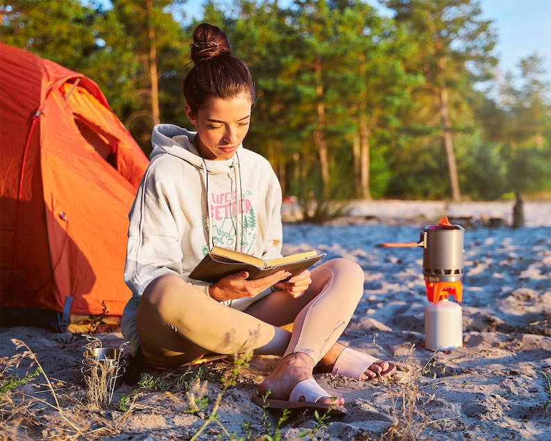 Pode incluir: Uma pessoa usando um moletom branco com a frase "LIFE IS BETTER" lendo um livro ao ar livre. Uma barraca laranja e um fog&atilde;o de acampamento s&atilde;o vis&iacute;veis ao fundo. A pessoa est&aacute; sentada na areia.