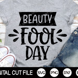 Op de afbeelding: Een grijs T-shirt met zwarte tekst die "Beauty Fool Day" luidt. De tekst is in een speels, handschriftlijk lettertype. Het shirt ligt plat op een houten oppervlak met een paar witte sneakers en een blauwe jeans op de achtergrond. De afbeelding bevat ook de tekst "DIGITAL CUT FILE SVG PNG DXF EPS".