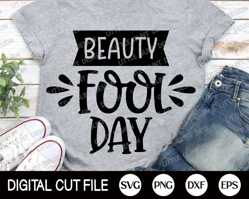 Op de afbeelding: Een grijs T-shirt met zwarte tekst die "Beauty Fool Day" luidt. De tekst is in een speels, handschriftlijk lettertype. Het shirt ligt plat op een houten oppervlak met een paar witte sneakers en een blauwe jeans op de achtergrond. De afbeelding bevat ook de tekst "DIGITAL CUT FILE SVG PNG DXF EPS".
