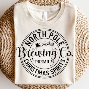 May include: A white t-shirt with a black circle design that says "North Pole Brewing Co. Est. 1824 Premium Christmas Spirits". The design also includes a silhouette of Santa Claus in a sleigh with reindeer.