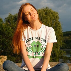 May include: A white t-shirt with a green four-leaf clover graphic and the text "Here for the Shenanigans".