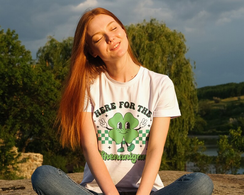 May include: A white t-shirt with a green four-leaf clover graphic and the text "Here for the Shenanigans".