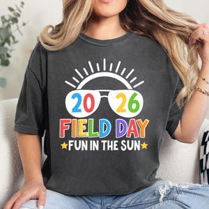 May include: Dark gray t-shirt with a colorful graphic. The design features the numbers "2026" in sunglasses, the words "FIELD DAY" in rainbow colors, and "FUN IN THE SUN" with star accents. The shirt has a casual, relaxed fit.