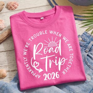 Pu&ograve; includere: Una maglietta rosa acceso con la scritta "Road Trip 2026" in un design circolare bianco. Il testo "Apparently we're trouble when we are together" circonda il testo principale. La maglietta &egrave; piegata su una superficie di legno chiaro.
