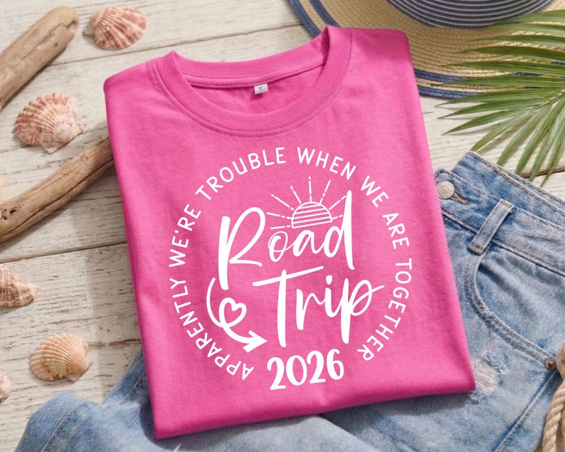 Pu&ograve; includere: Una maglietta rosa acceso con la scritta "Road Trip 2026" in un design circolare bianco. Il testo "Apparently we're trouble when we are together" circonda il testo principale. La maglietta &egrave; piegata su una superficie di legno chiaro.