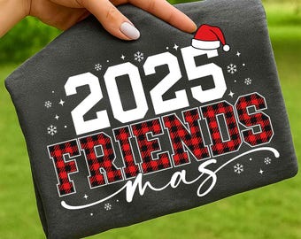 Op de afbeelding: Donkergrijs T-shirt met de tekst "2025 FRIENDS mas" in wit en rood geruit. Een kerstmuts-afbeelding staat boven het jaar en sneeuwvlokken zijn verspreid rond de tekst. Het T-shirt wordt door een hand vastgehouden.