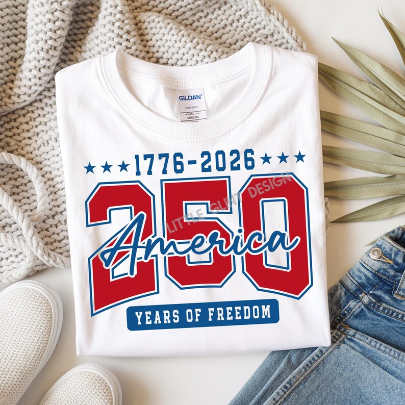 May include: White t-shirt with a patriotic design celebrating America's 250th anniversary. The design features the numbers "250" in red, the word "America" in blue script, and the years "1776-2026" with stars. The text "YEARS OF FREEDOM" is also included.