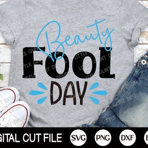May include: A gray t-shirt with the text "Beauty Fool Day" in black and blue lettering. The text "Fool" is in large, bold letters. The text "Beauty" is in a cursive font. The text "Day" is in a smaller font and has blue water droplets around it.