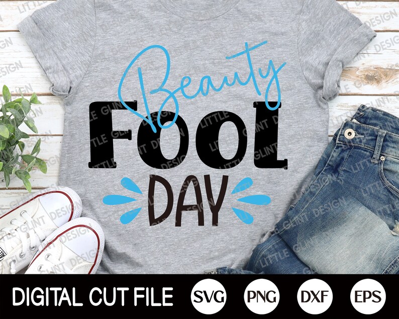 May include: A gray t-shirt with the text "Beauty Fool Day" in black and blue lettering. The text "Fool" is in large, bold letters. The text "Beauty" is in a cursive font. The text "Day" is in a smaller font and has blue water droplets around it.