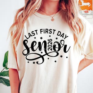 May include: Cream-colored t-shirt with the text "LAST FIRST DAY SENIOR 2026" in black script and block letters, with star accents. The shirt is a casual style.