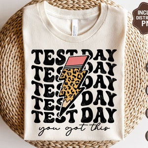 May include: A white t-shirt with the text "TEST DAY" repeated in a retro font, with a leopard print lightning bolt graphic and the text "you got this".