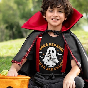 May include: A child dressed as a vampire for Halloween is wearing a black t-shirt with a white ghost and a dog skeleton inside. The shirt says "Dogs Because People Are Creepy".