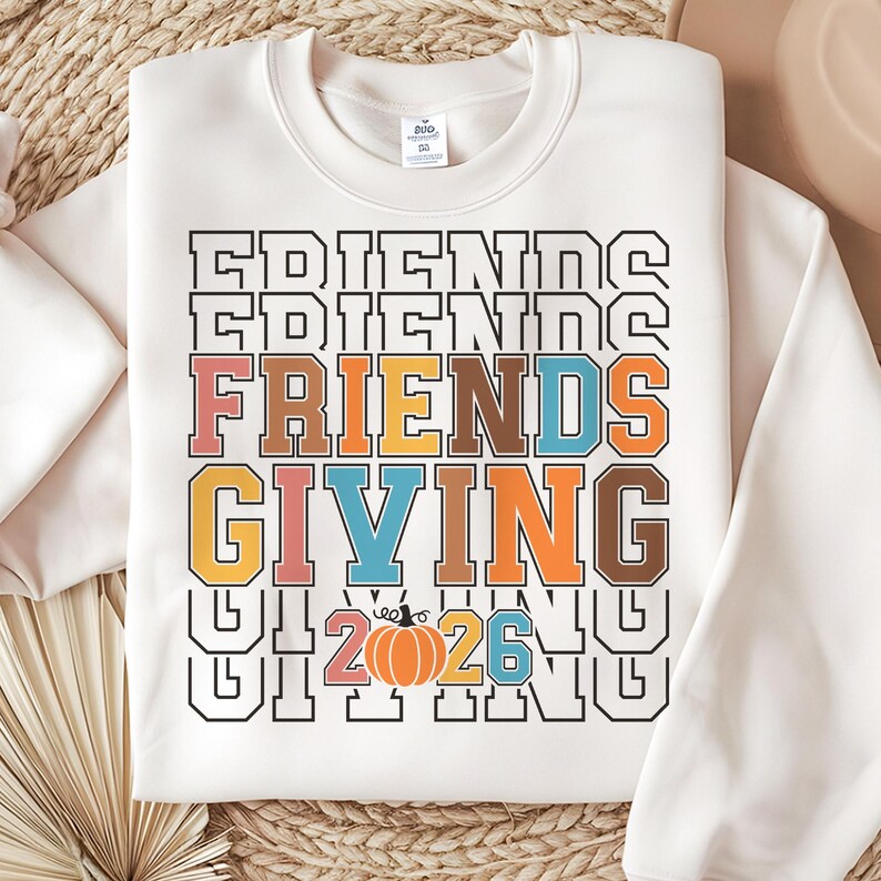 May include: A cream-colored crewneck sweatshirt featuring a multi-layered graphic print. The text reads FRIENDS GIVING in a collegiate font with colors including orange, blue, yellow, and brown. A small orange pumpkin replaces the zero in 2026.