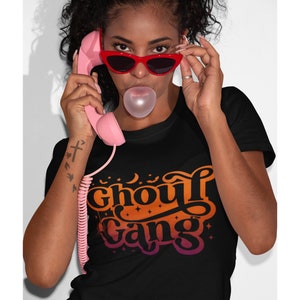 May include: A black t-shirt with an orange, yellow, and pink gradient design that says "Ghoul Gang" with stars and bats around the text.