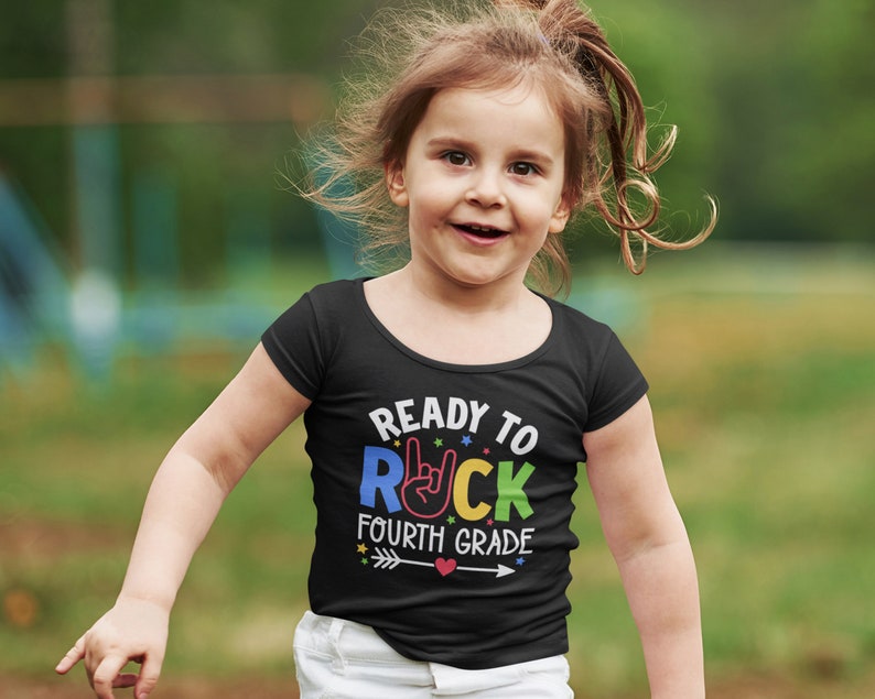 May include: A young person wearing a black t-shirt with the text "Ready to Rock Fourth Grade" in rainbow colors. The text includes a rainbow colored rock and roll hand gesture.