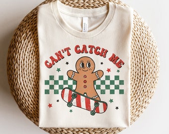 Op de afbeelding: Een beige T-shirt met een peperkoekman op een skateboard. De peperkoekman draagt een rood-wit gestreepte muts en heeft een rood-wit gestreept skateboard. De tekst "Can't Catch Me" staat in rood-witte letters boven de peperkoekman.