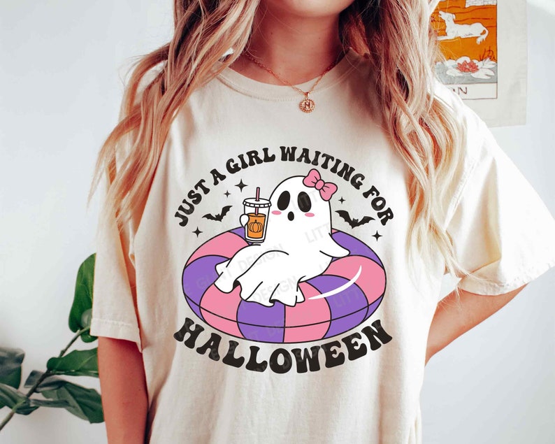Może przedstawiać: Biały T-shirt z grafiką przedstawiającą ducha w r&oacute;żowej kokardce trzymającego latte z przyprawą dyniową w r&oacute;żowym kubku. Duch siedzi w fioletowo-r&oacute;żowym dmuchanym k&oacute;łku. Na koszulce nadrukowany jest czarny napis "Just a girl waiting for Halloween".