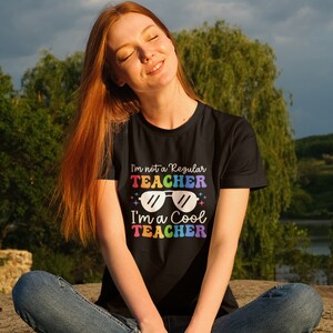 May include: A woman wearing a black t-shirt with the text "I'm not a regular teacher I'm a cool teacher" in rainbow colours and sunglasses graphic.