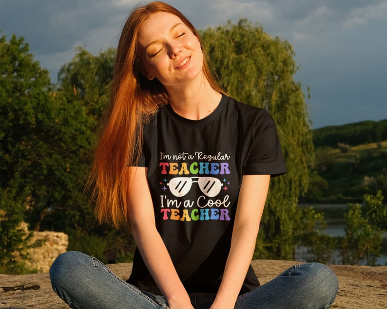 May include: A woman wearing a black t-shirt with the text "I'm not a regular teacher I'm a cool teacher" in rainbow colours and sunglasses graphic.