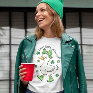 May include: White t-shirt with a graphic of a goose wearing a green bow and boots, surrounded by shamrocks. The text "Lucky Goose on the Loose" is above the goose. The person is wearing a green jacket and hat, holding a red cup.
