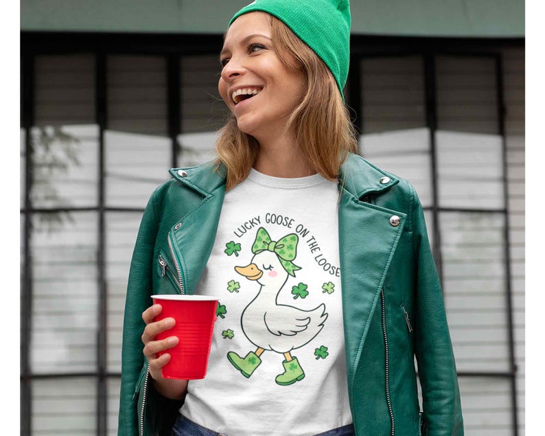 May include: White t-shirt with a graphic of a goose wearing a green bow and boots, surrounded by shamrocks. The text "Lucky Goose on the Loose" is above the goose. The person is wearing a green jacket and hat, holding a red cup.