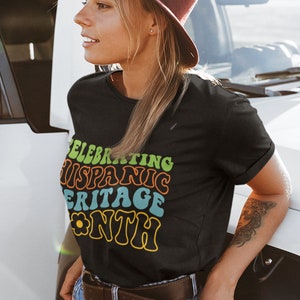 May include: A black t-shirt with a colorful graphic that says "Celebrating Hispanic Heritage Month" in a retro font. The graphic includes a yellow flower.